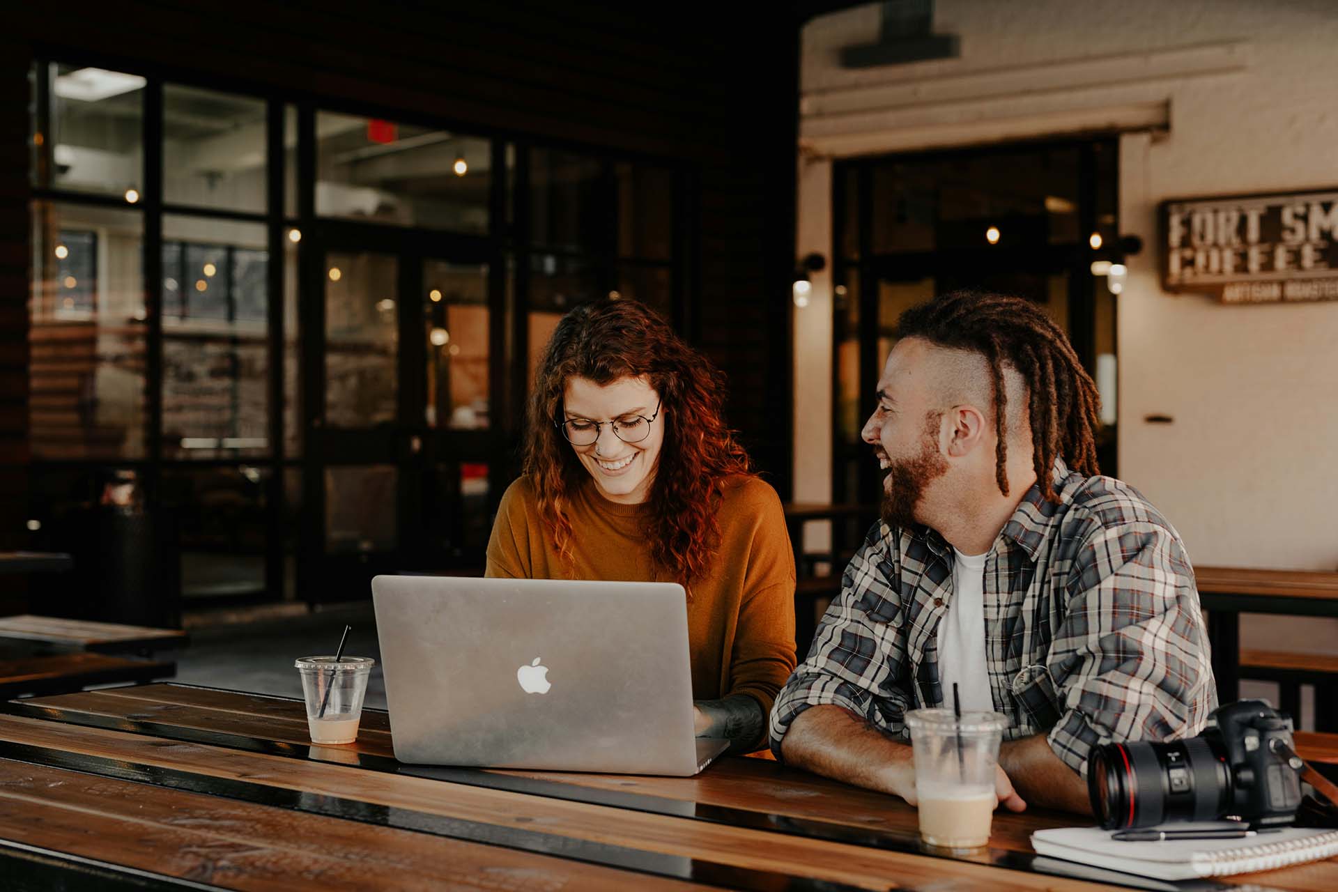 Two friends smiling and laughing working together on computer at cafe.